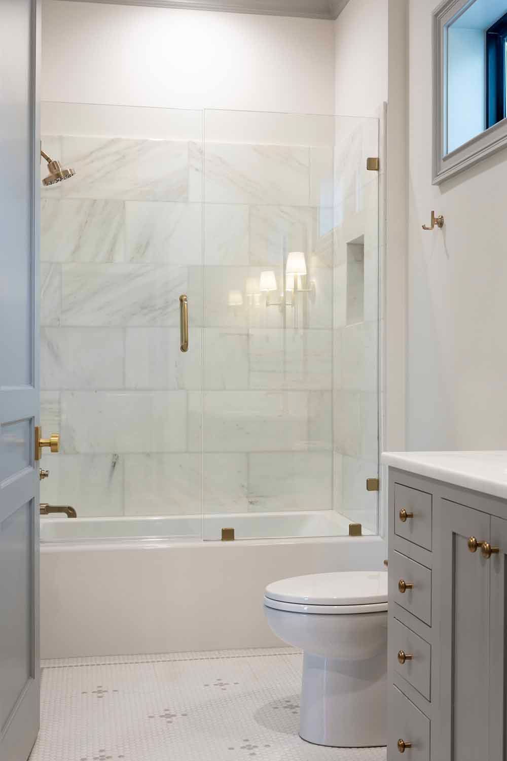 A modern bathroom featuring a white marble-tiled bathtub with a glass door, a white toilet, and a grey vanity cabinet.