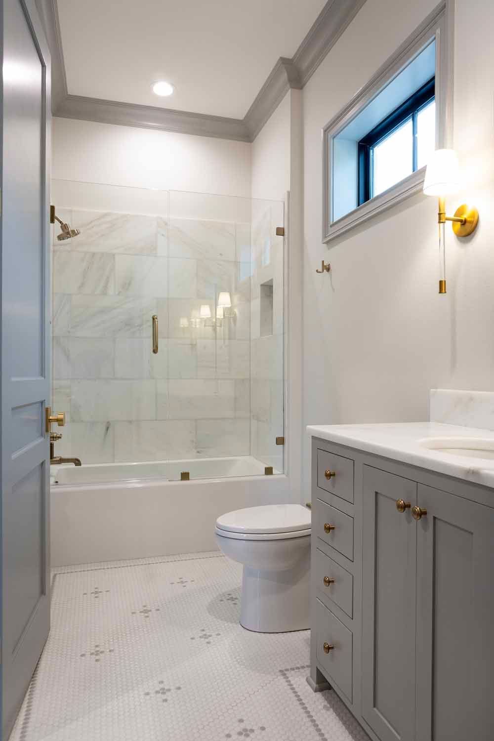 A bright, modern bathroom featuring a grey vanity, white marble shower, patterned tile floors, and gold hardware.