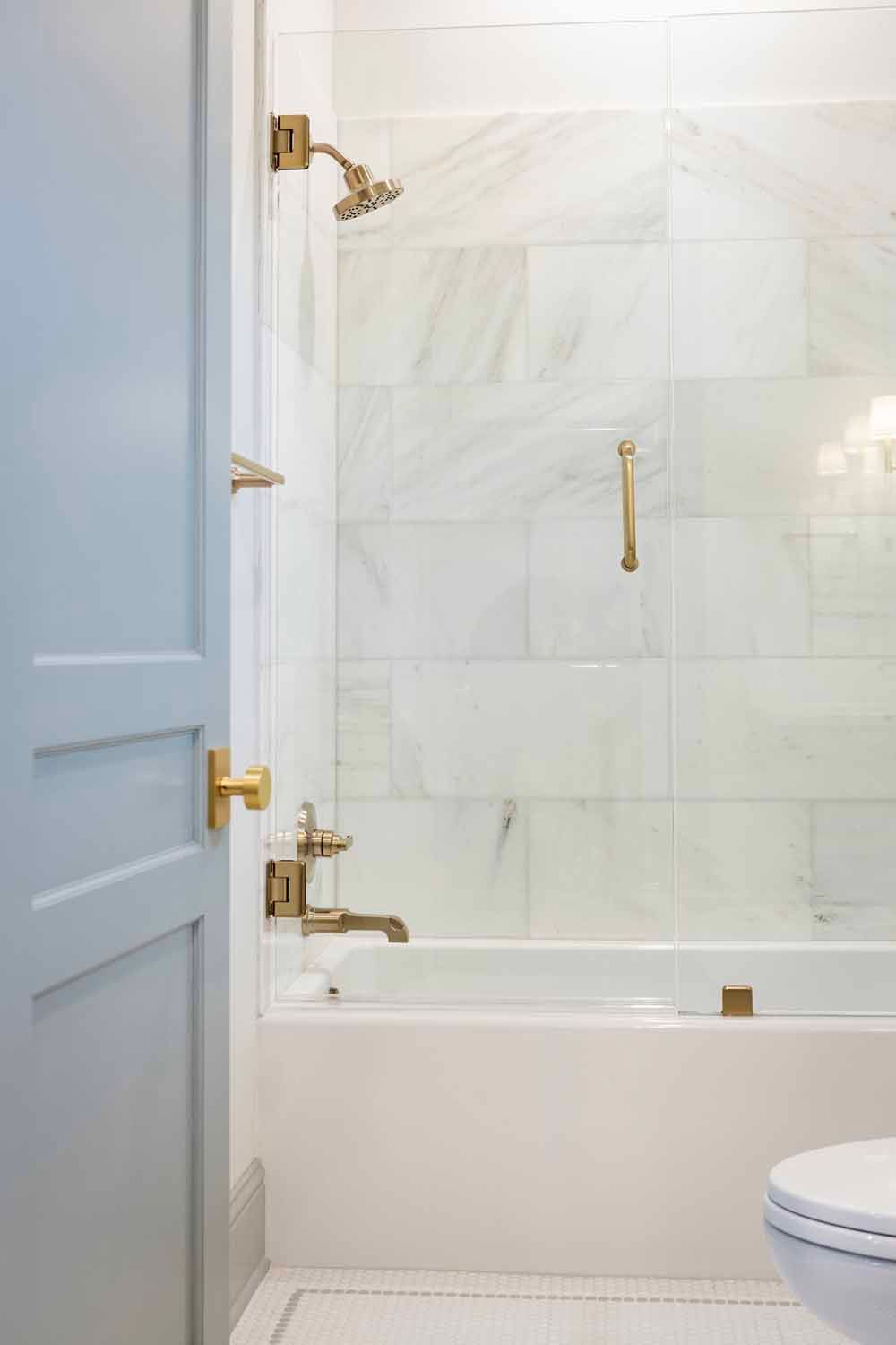 A light blue door opens to a bathroom with a white bathtub, marble tile walls, and brass hardware.
