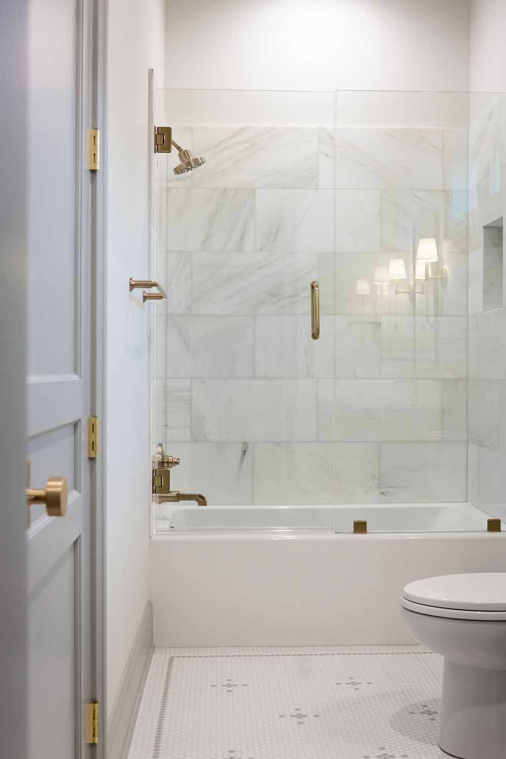 A white marble-tiled bathtub and shower with a glass panel, gold fixtures, and a white tiled floor in a bathroom.