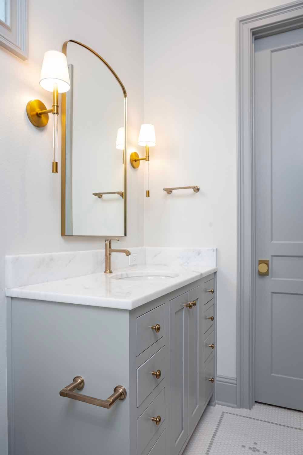 A modern bathroom vanity with a gray cabinet, marble countertop, brass hardware, wall sconce lighting, and arched mirror.