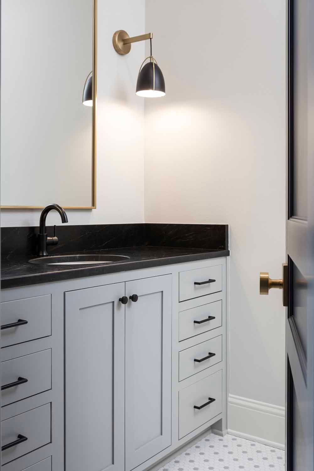 A bathroom vanity with a light gray cabinet, black marble countertop, brass sconce, and small black-and-white floor tiles.