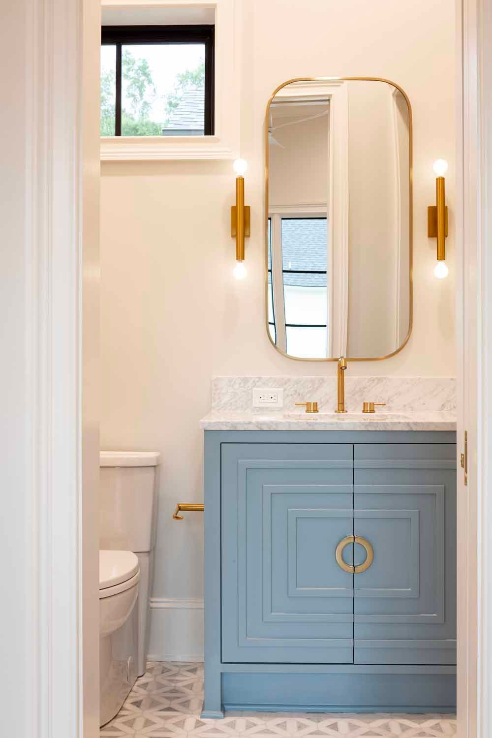 A modern powder room with a blue, geometric-patterned vanity, gold hardware, and an arched mirror under a small window.