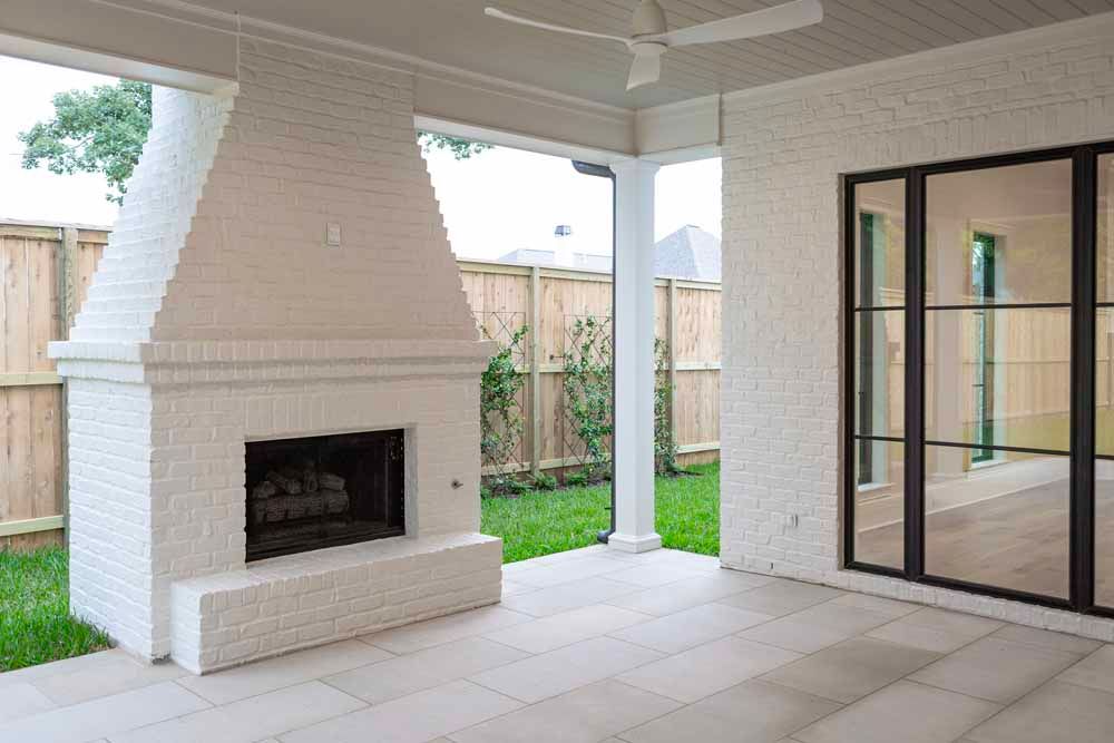 A modern white brick outdoor fireplace on a patio next to large glass doors and a wooden fence.