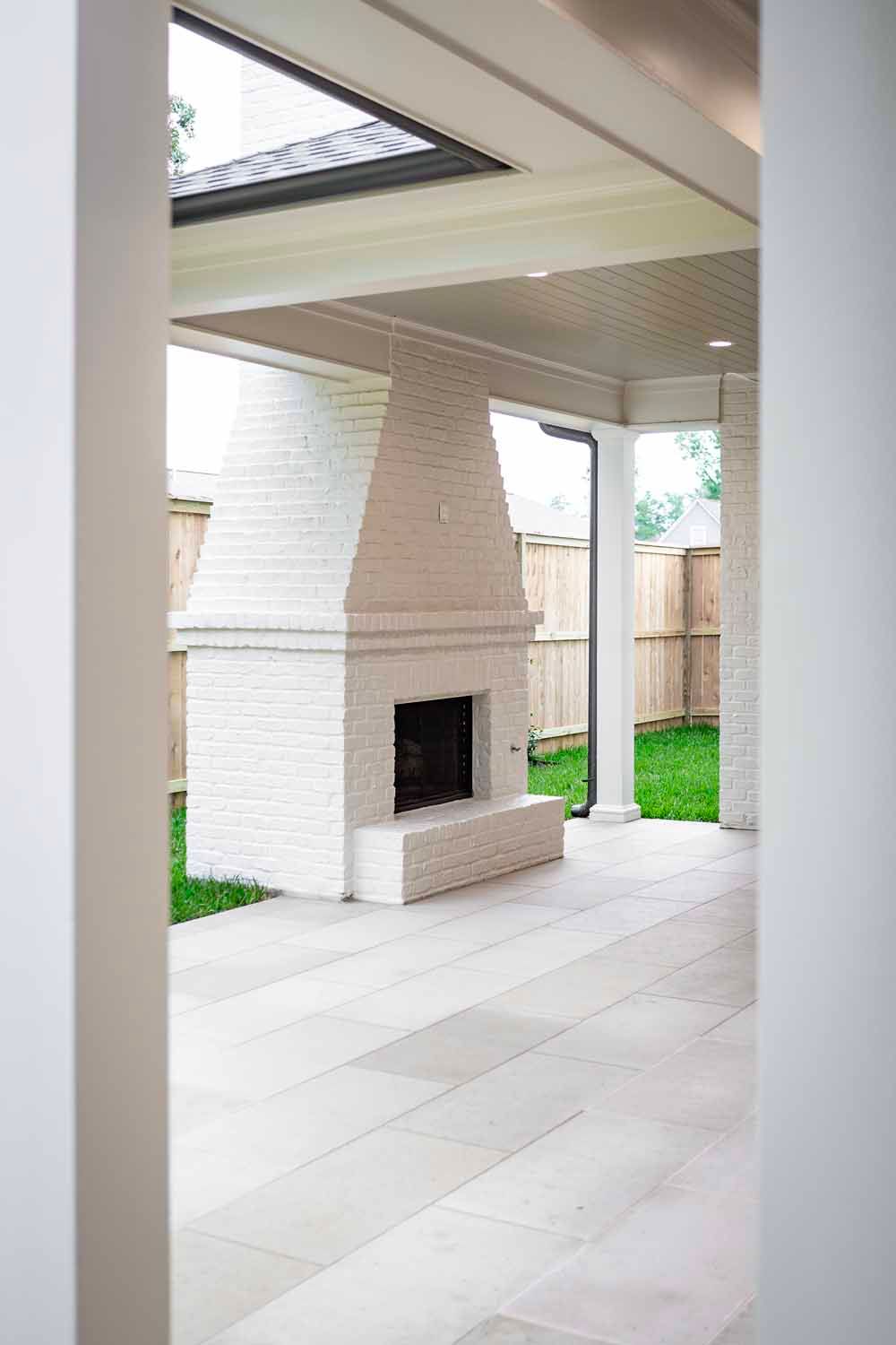 A covered outdoor patio featuring a white brick fireplace, light stone flooring, and a view of the grassy yard.