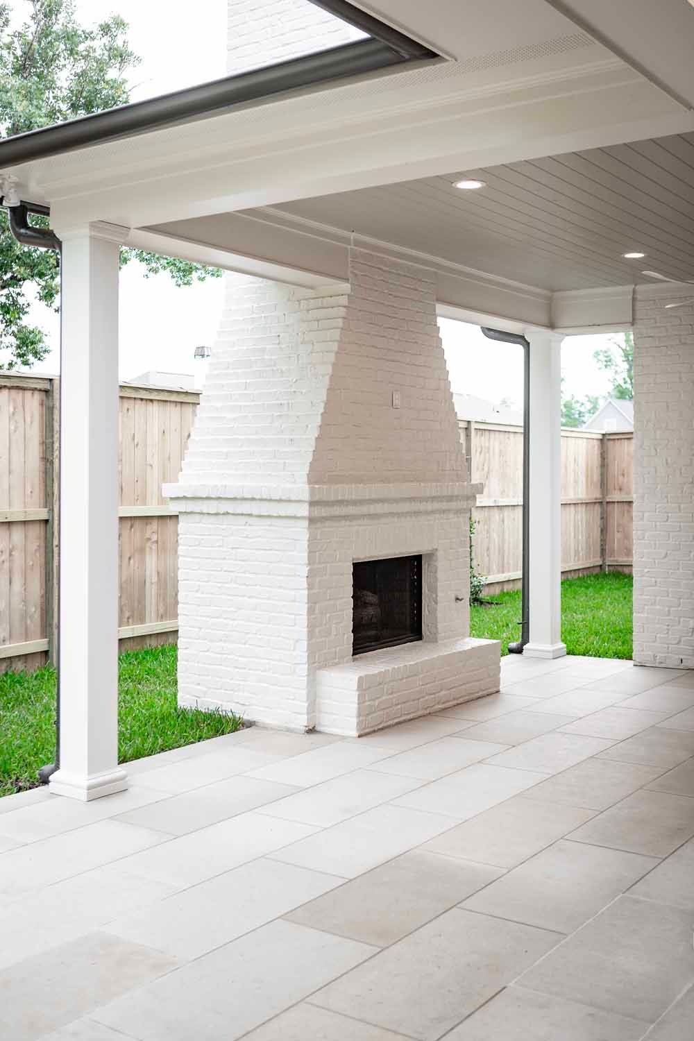 A white brick outdoor fireplace stands on a patio under a covered porch with stone flooring and a wooden privacy fence.