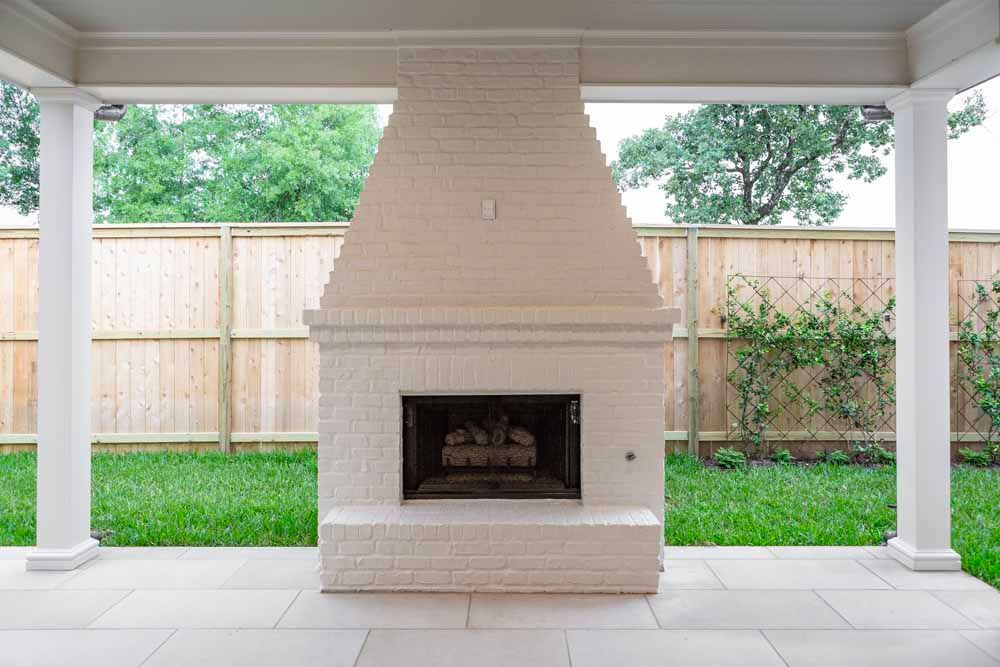 A beige painted brick outdoor fireplace centered under a covered patio with a wooden fence and lawn in the background.