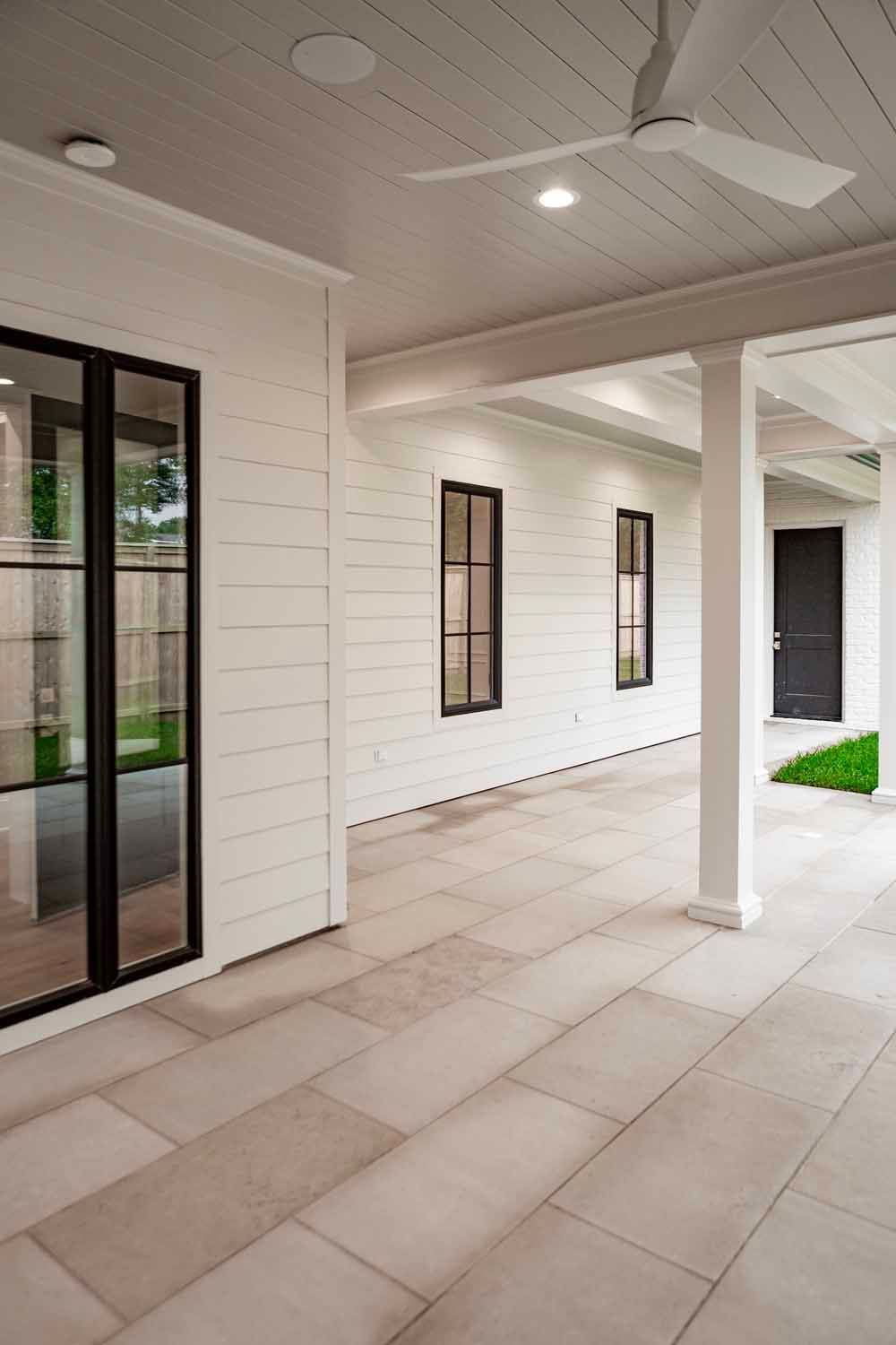 A white, modern covered porch with horizontal siding, large black-framed windows, a ceiling fan, and light stone flooring.