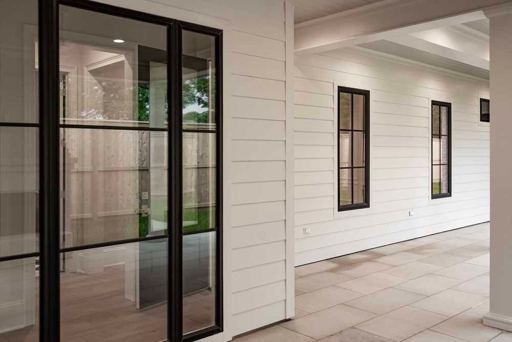 White lap-sided exterior wall with black-framed windows and a glass door opening onto a paved porch.