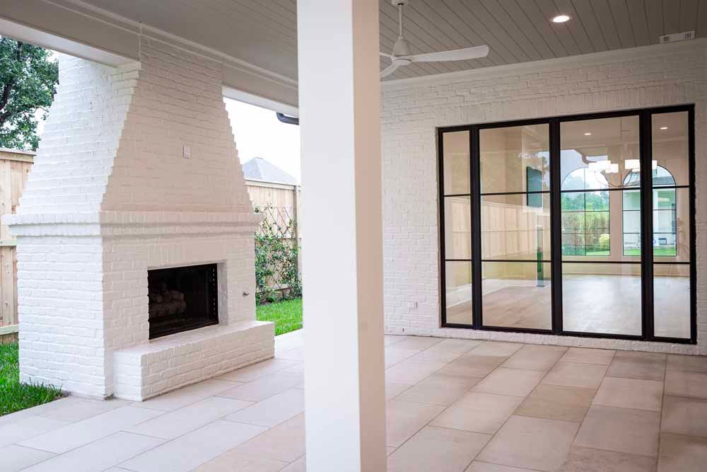 A white-bricked outdoor patio featuring a fireplace, a ceiling fan, and large black-framed glass sliding doors.