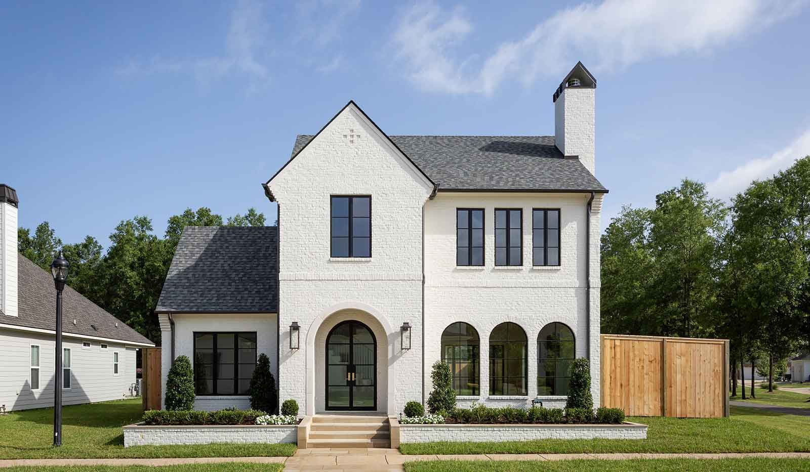 Two-story white brick house with a dark gabled roof, an arched front entryway, and a chimney on a sunny, grassy lot.
