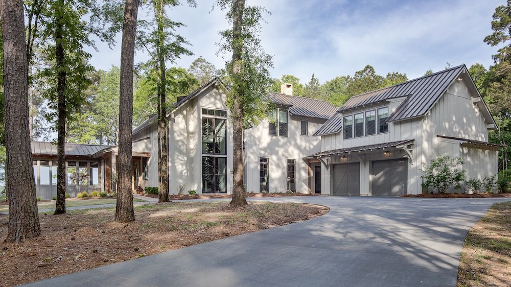 A two-story, white modern house with a gray metal roof, attached garage, and surrounding trees, set along a driveway.