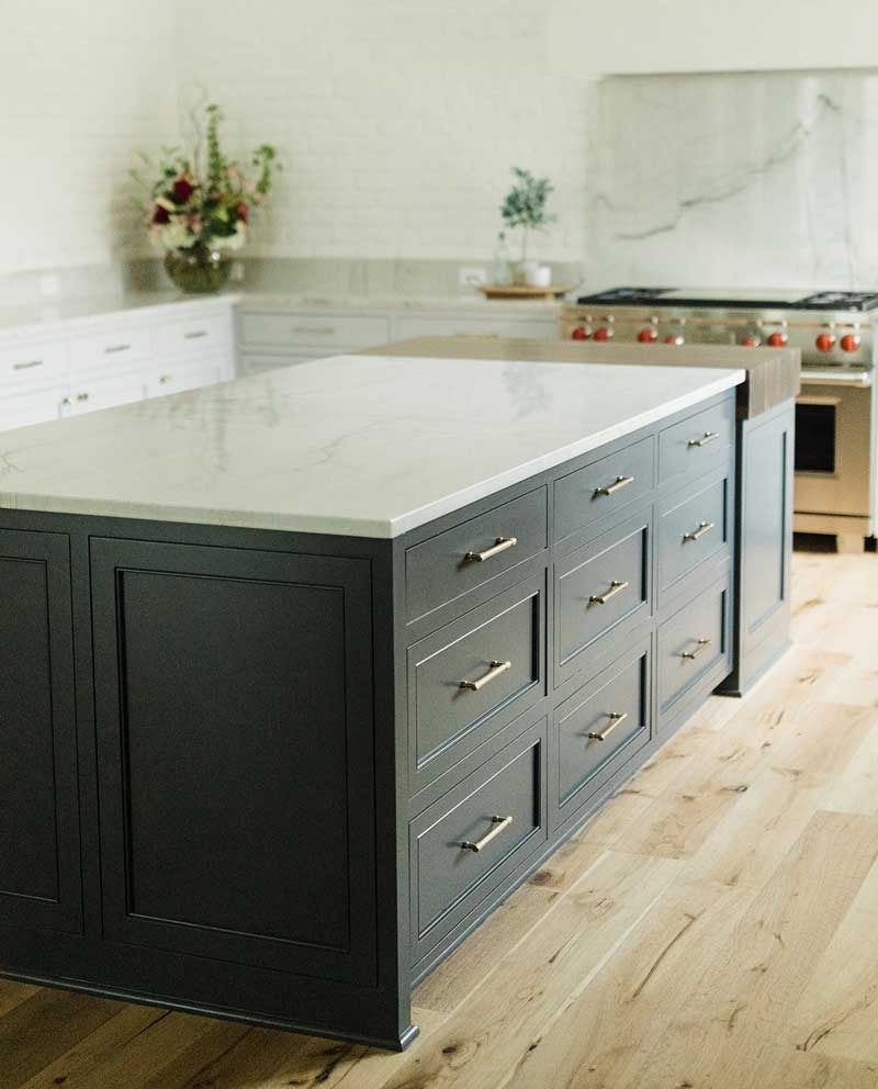 A large, dark kitchen island with a white marble countertop stands in a bright kitchen with light wooden floors.