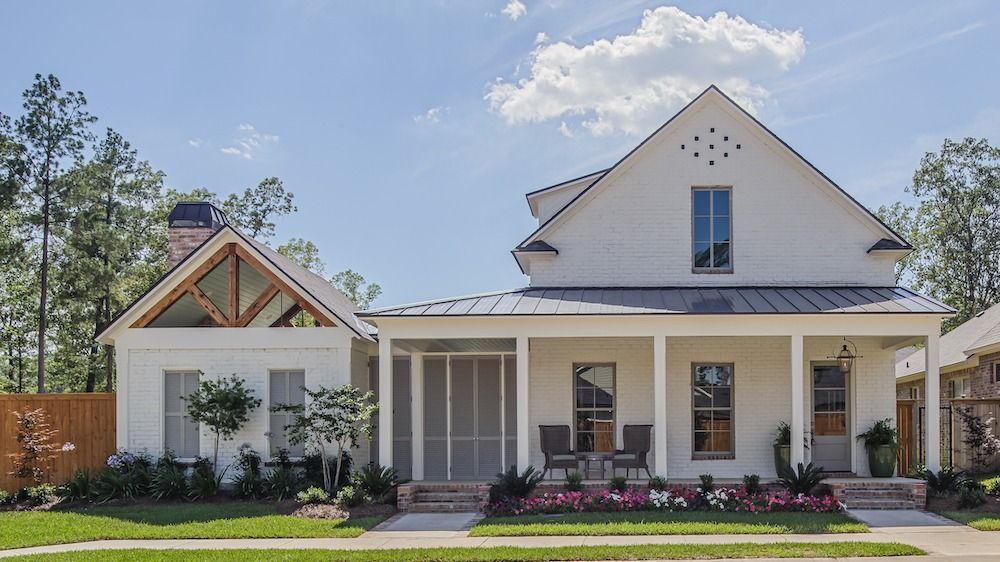 A white farmhouse with a metal roof, front porch, and screened-in area, surrounded by a lawn and trees under a blue sky.