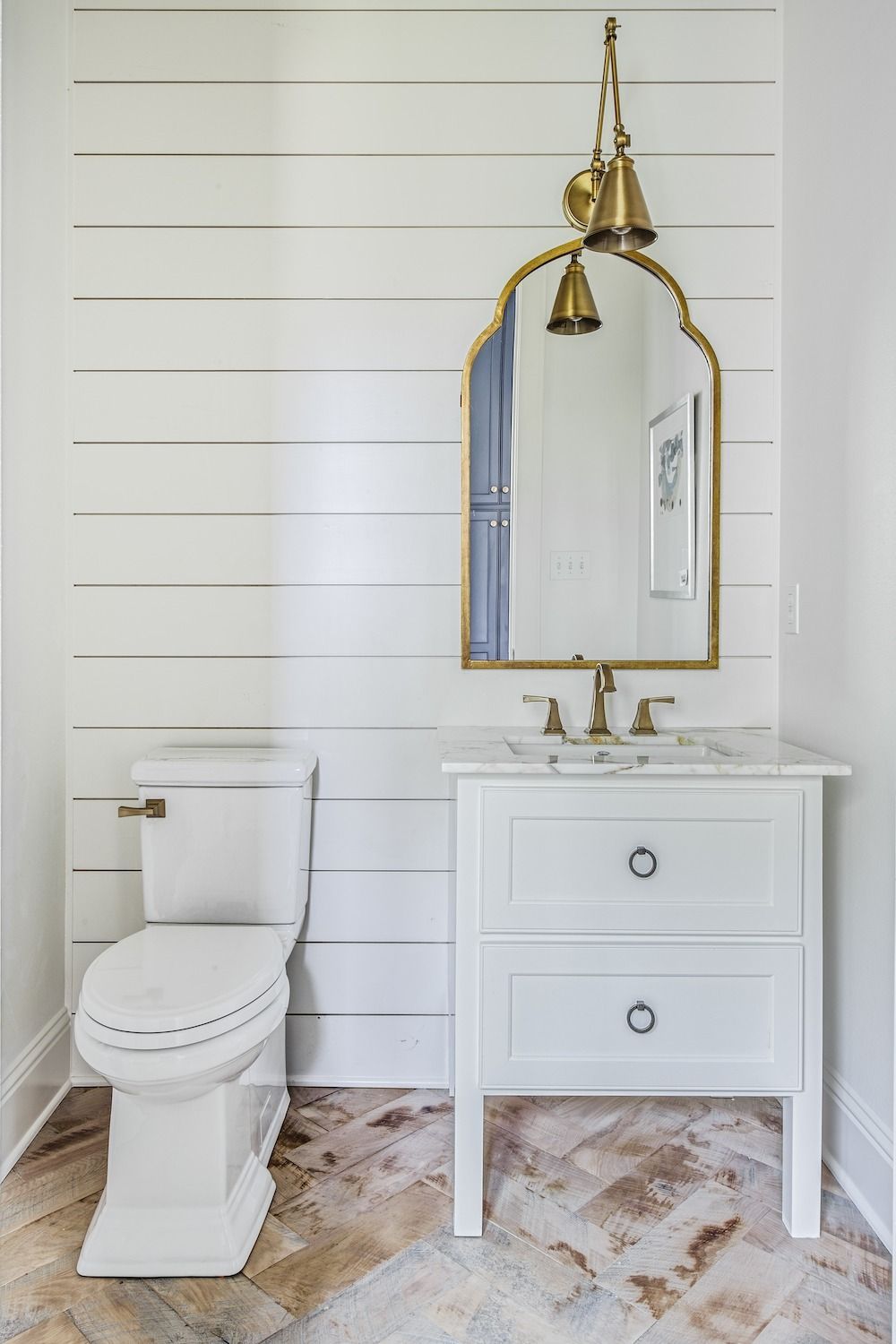 A white bathroom with shiplap walls, a wooden vanity, a gold-framed arched mirror, and patterned tile flooring.