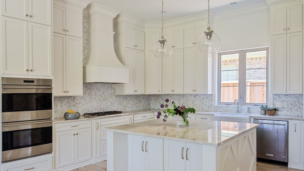 A modern white kitchen featuring a large island, stainless steel appliances, marble backsplash, and hanging pendant lights.