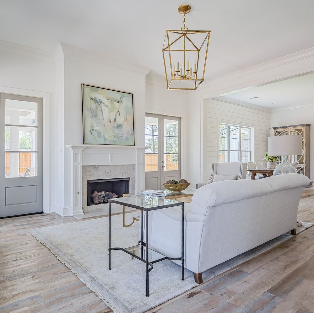 A bright living room with a white sofa, glass end table, fireplace, framed art, and a gold geometric pendant light.