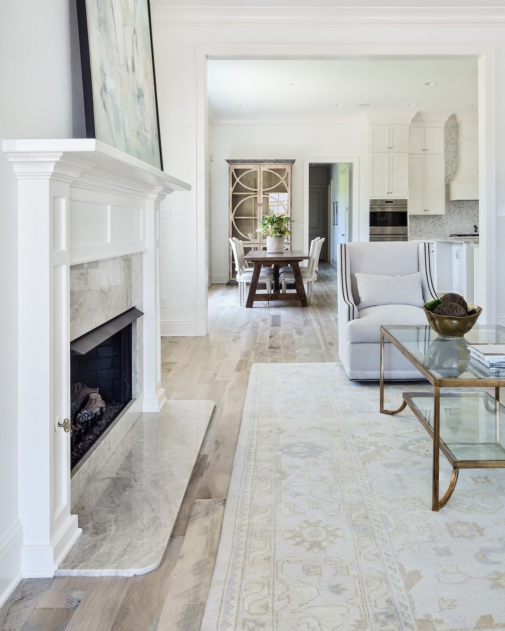 A living room with a white fireplace, light wood floors, a neutral rug, and a clear coffee table looking into a dining area.