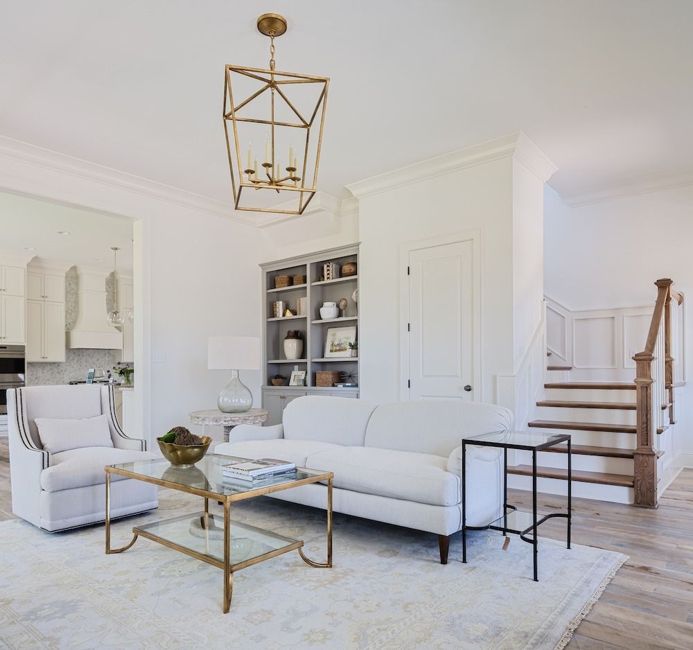 A bright living room with a white sofa, armchair, glass coffee table, and gold lantern light, with a staircase nearby.