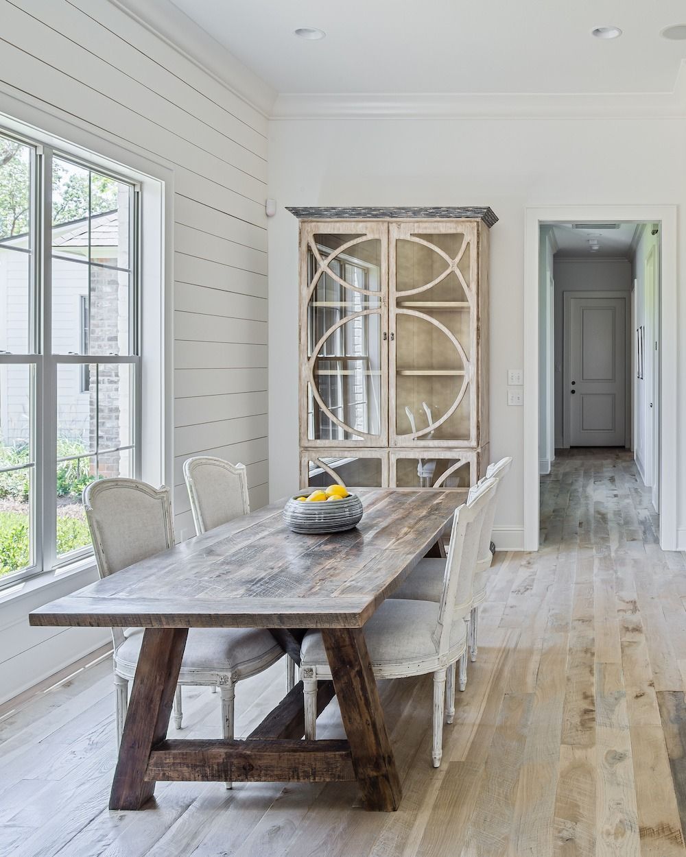 A rustic wooden dining table with four cushioned chairs sits in a bright room with shiplap walls and a wooden cabinet.