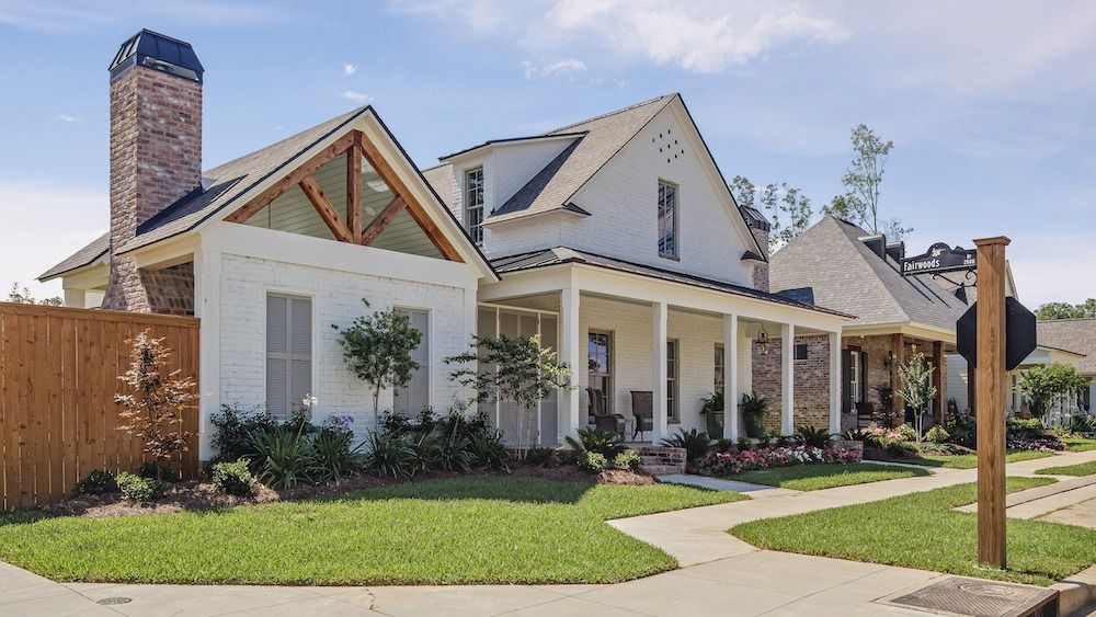 A white, farmhouse-style home with a brick chimney and front porch sits on a grassy lot under a blue sky.
