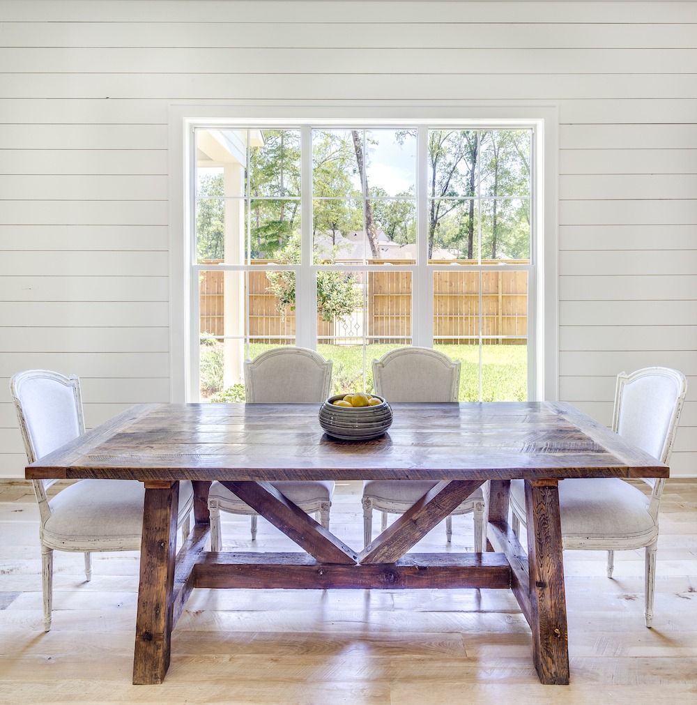 A rustic wooden dining table with four white upholstered chairs in a room with white shiplap walls and a large window.