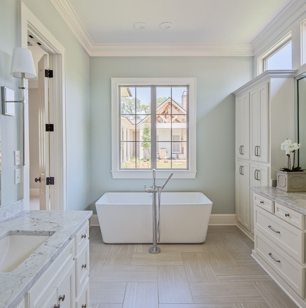 A serene, light-blue bathroom with a freestanding white tub, dual marble vanities, and bright white cabinetry.