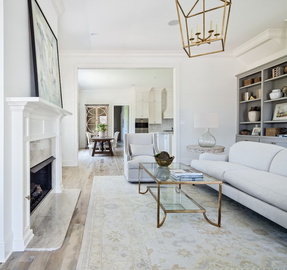 A bright, elegant living room with a fireplace, white sofa, glass coffee table, and open transition to a dining area.
