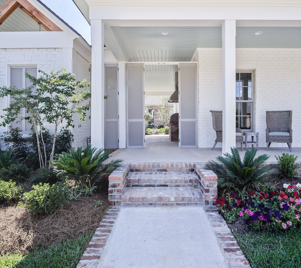 A front porch with white brick, a light blue ceiling, a concrete walkway, brick steps, two wicker chairs, and greenery.