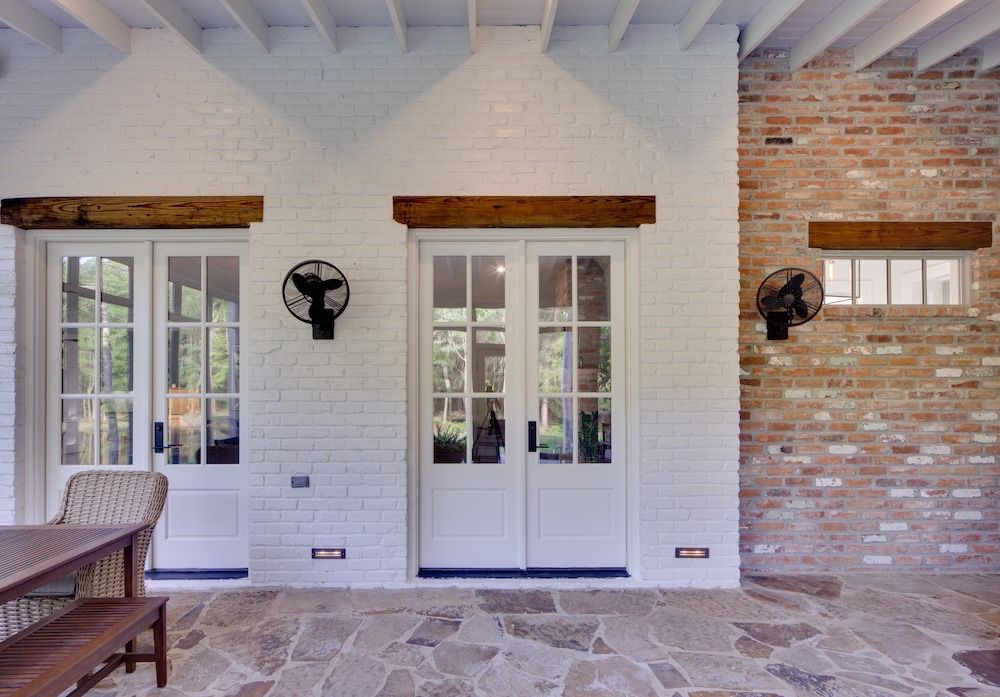 A covered patio with a stone floor, featuring two sets of white French doors, rustic wood beams, and a brick wall.