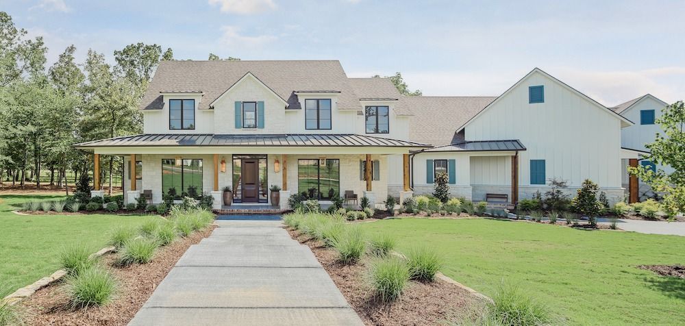 A two-story white farmhouse with a metal-roofed porch, blue shutters, and a concrete driveway in a grassy, wooded setting.
