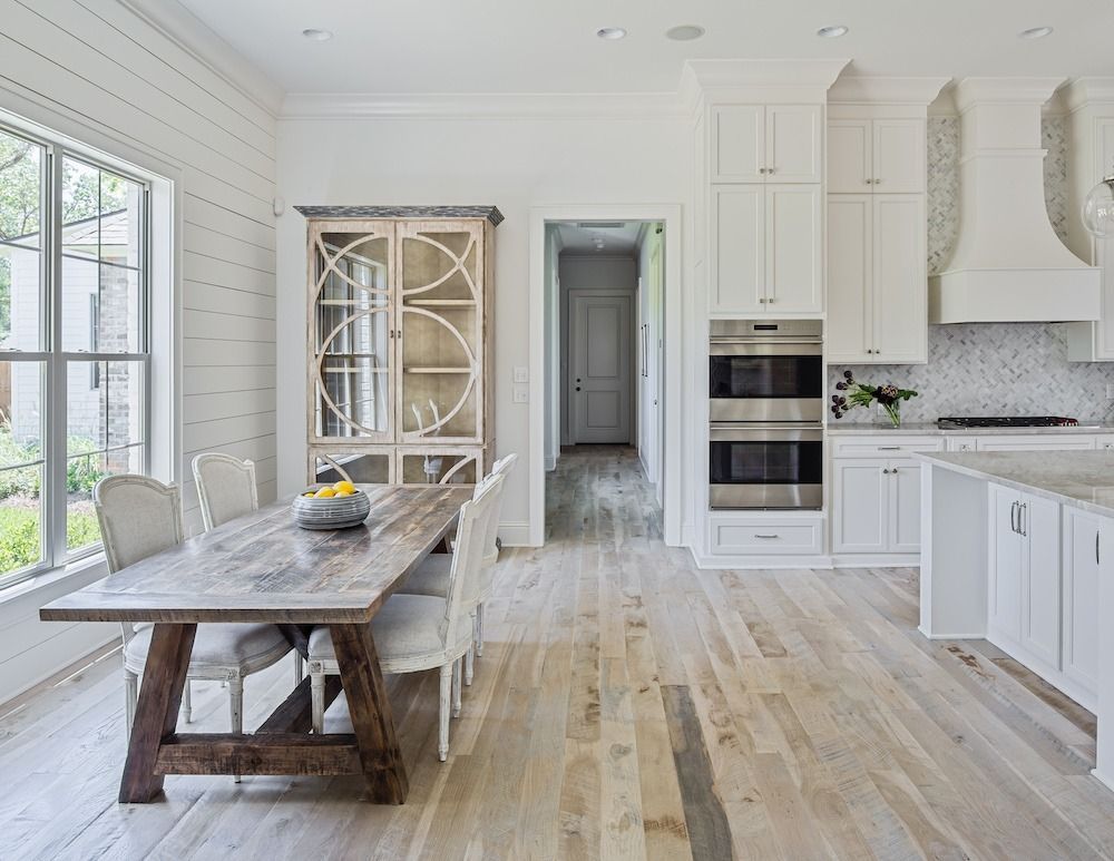 A bright kitchen and dining area featuring white cabinets, a wooden dining table, a glass cabinet, and light wood flooring.