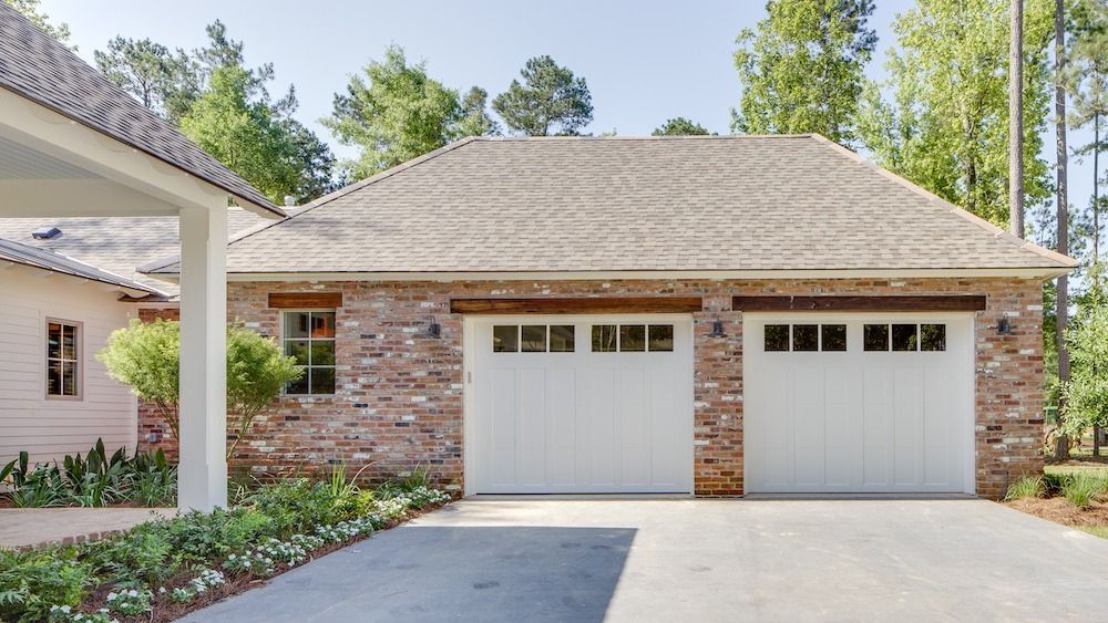 A brick two-car garage with white carriage-style doors and a light gray shingled roof, surrounded by trees and landscaping.