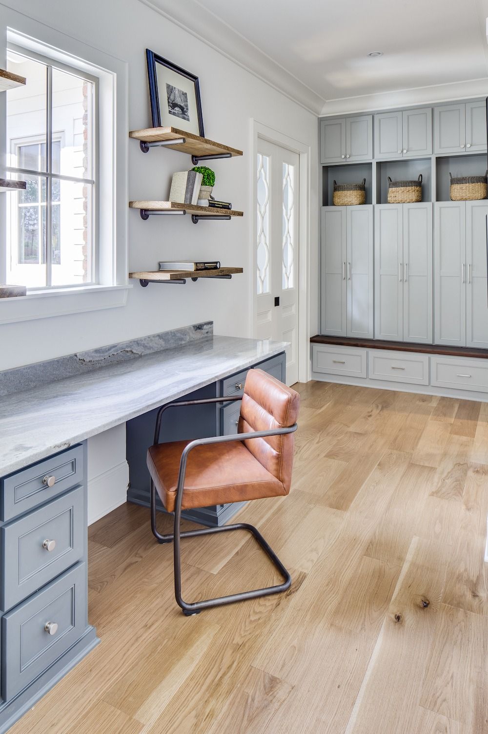 A desk with a leather chair, open shelving, and light gray cabinetry in a sunlit home office with light wood floors.