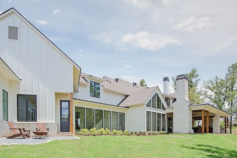 White modern farmhouse with vertical siding, a stone chimney, a covered patio, and large windows on a green lawn.