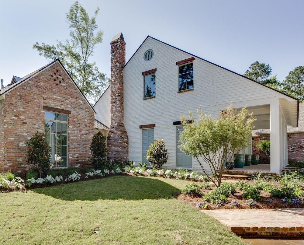 A white house with a tall brick chimney, stone accent wall, large windows, and a landscaped front lawn on a sunny day.