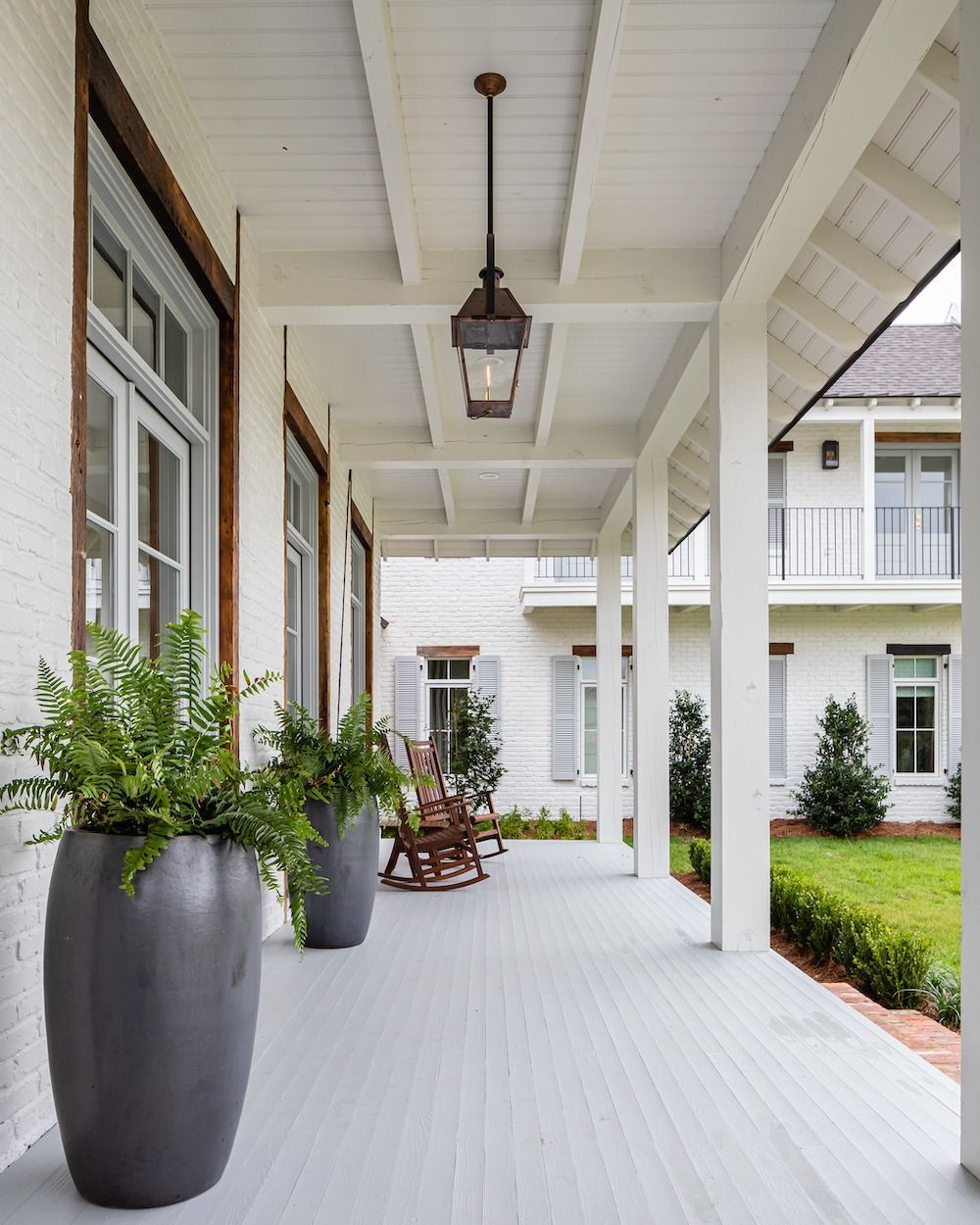 A covered porch with white wooden pillars, grey flooring, large dark planters, and hanging lanterns overlooking a lawn.