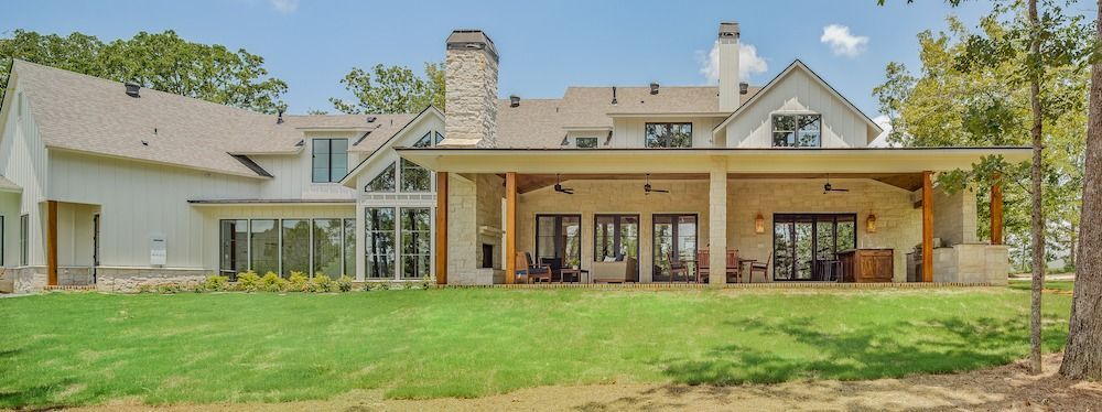 A large, light-colored modern farmhouse with a stone fireplace and a covered back patio overlooking a grassy lawn.