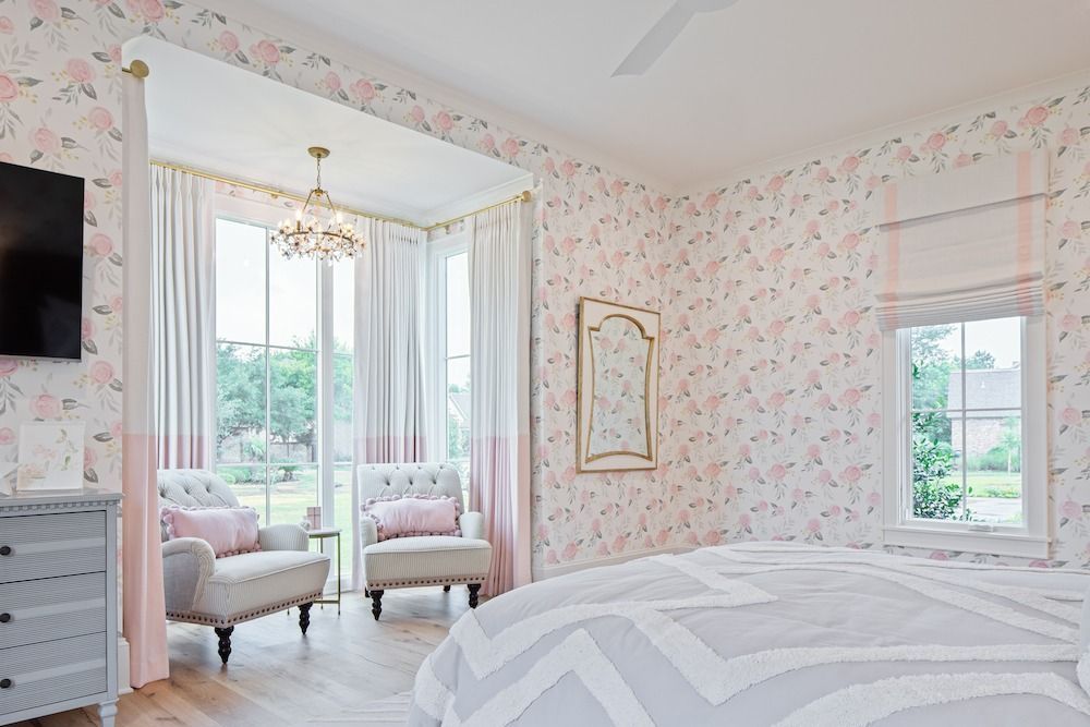 A bright bedroom features floral wallpaper, a bed with a chevron quilt, and a seating nook with two chairs by a window.