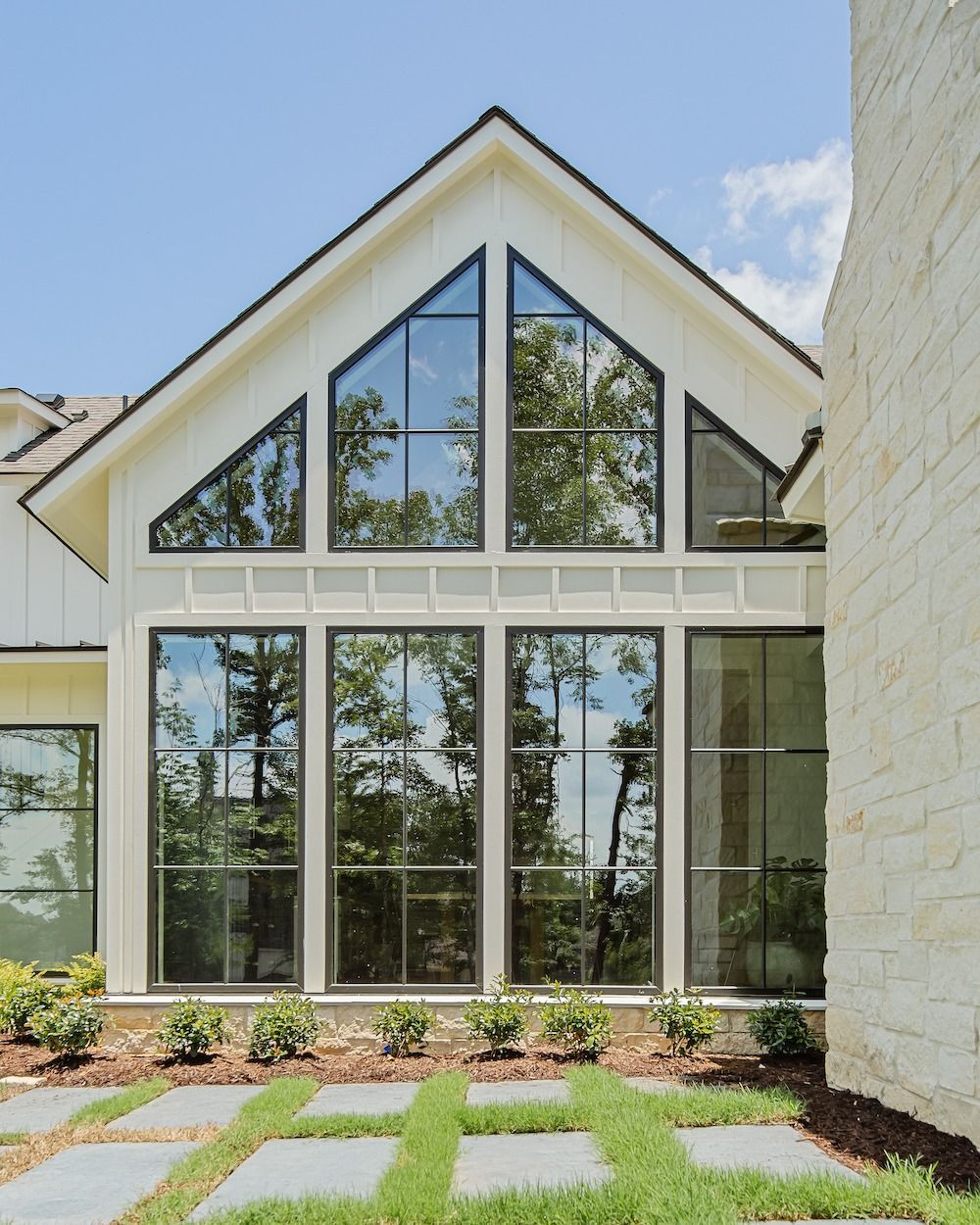 A white, gable-roofed house features large, grid-patterned windows overlooking a patio with stone pavers and green grass.