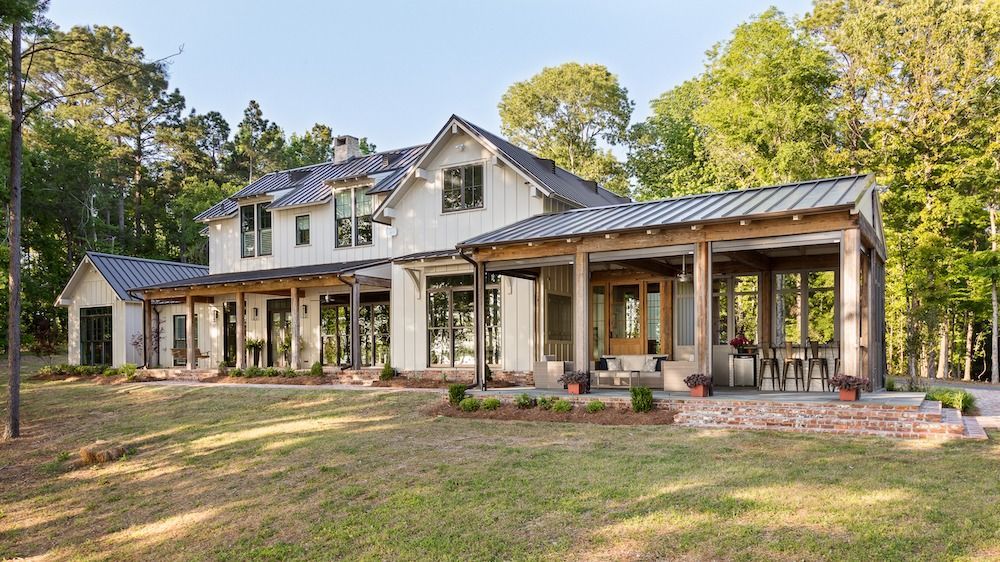A two-story white farmhouse with a dark metal roof, large windows, and an expansive covered porch set in a wooded area.