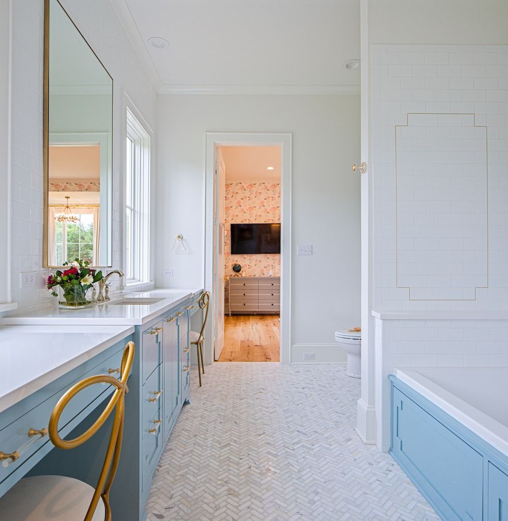 A bright bathroom with light blue cabinets, a gold-framed mirror, and white herringbone tile flooring.