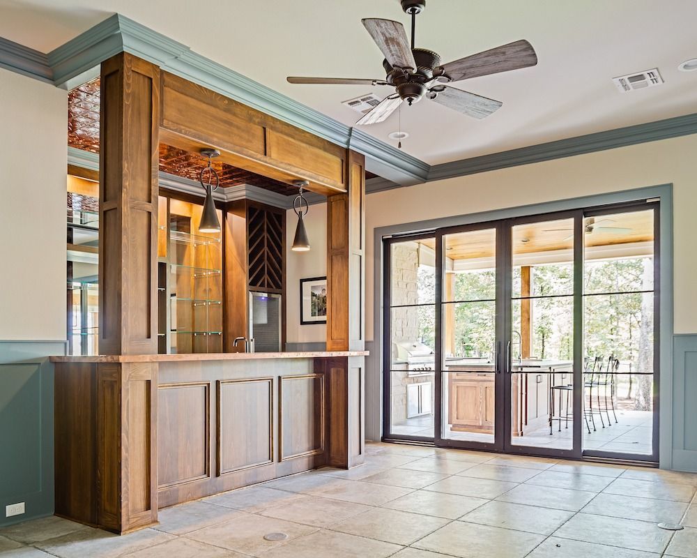 A wooden home bar with glass shelving and an integrated wine rack, situated in a room with a ceiling fan and patio doors.