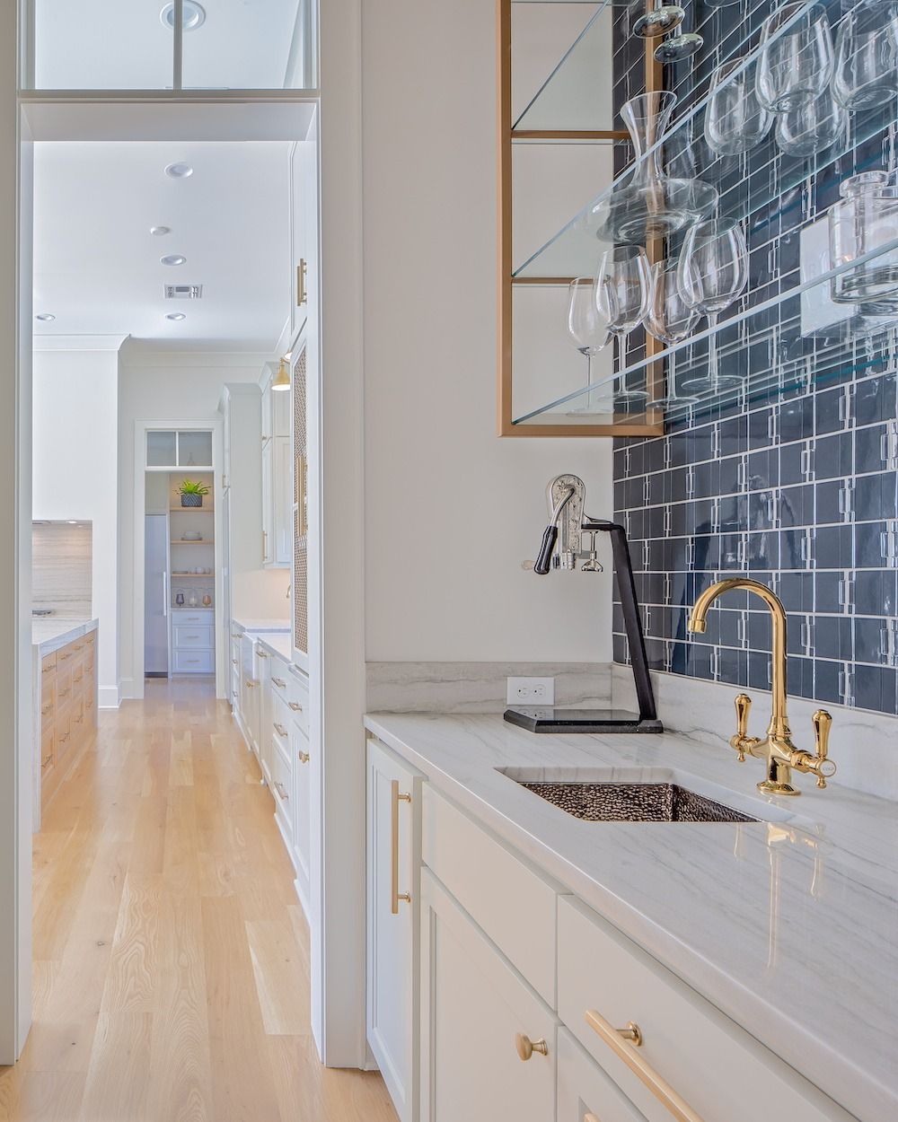 A bright kitchen wet bar with a gold faucet, white marble countertop, dark patterned tile backsplash, and wooden floors.