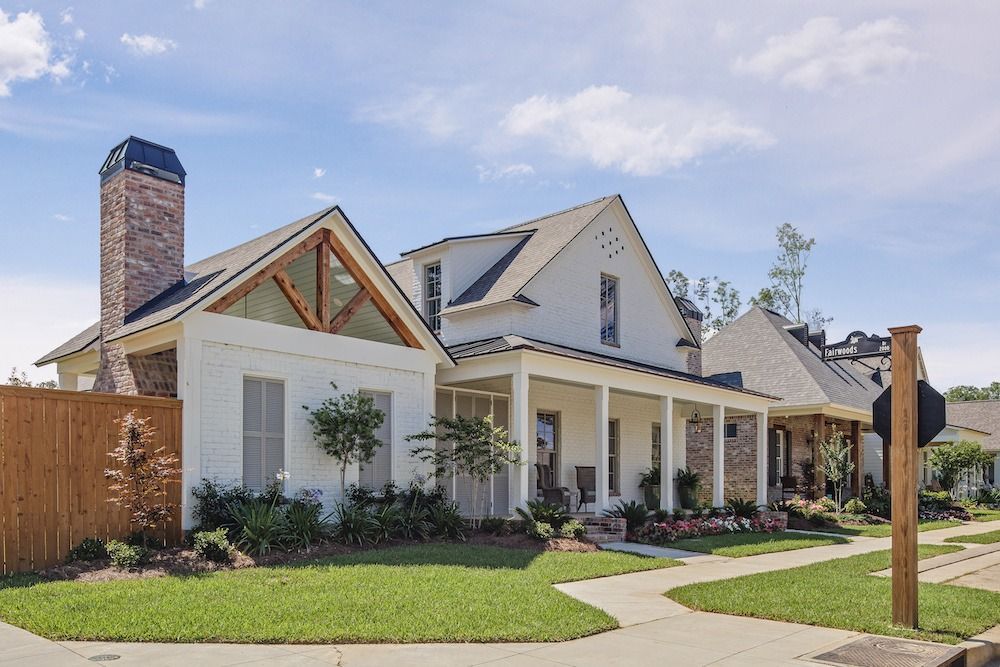 A white cottage-style house with a brick chimney and a wooden porch, situated in a suburban neighborhood on a sunny day.