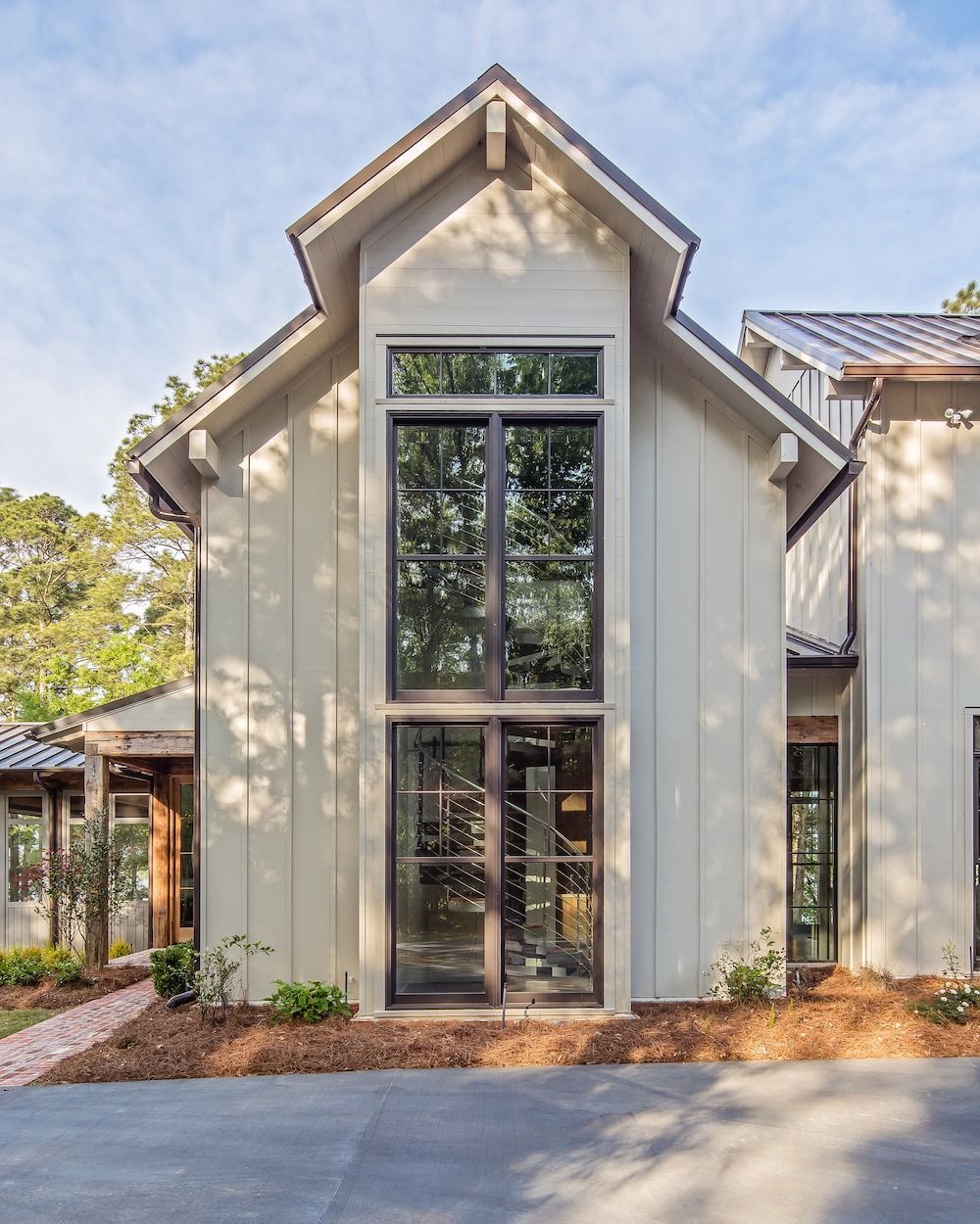 A modern home exterior with cream-colored vertical siding, dark window frames, a gabled roof, and a landscaped front yard.