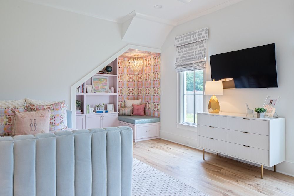 A bedroom with a pale blue tufted bed, a white dresser with a television, and a cozy built-in reading nook with wallpaper.
