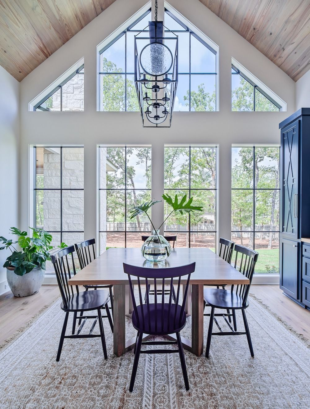 A sunlit dining room with a wooden table, black spindle chairs, a large lantern light, and floor-to-ceiling windows.