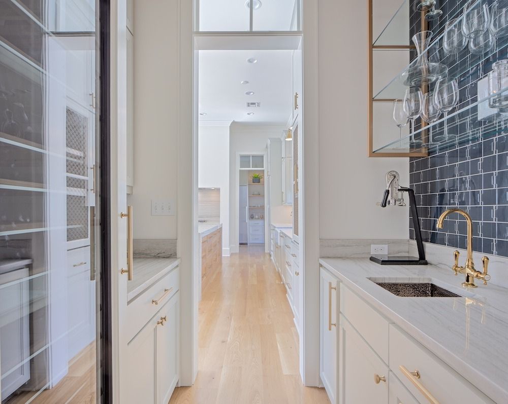 A bright butler's pantry with white cabinets, marble countertops, a gold faucet, and a black tiled wall leading to a kitchen.