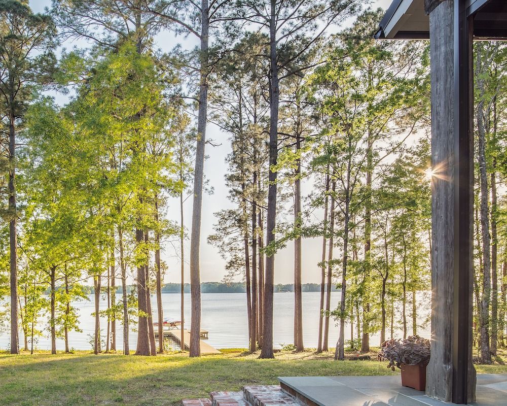 A sunny lakeside view framed by tall pine trees, seen from a stone patio with a wooden post in the foreground.